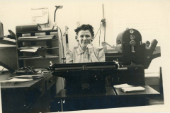 Helen Law Manzer at typewriter with mimeograph machine behind her.