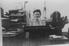 Helen Law Manzer at typewriter with mimeograph machine behind her.