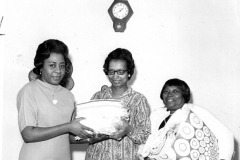 Three women under a clock, two holding a folded cloths with a third holding a bag filled with them.