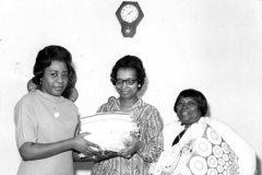 Three women under a clock, two holding a folded cloths with a third holding a bag filled with them.